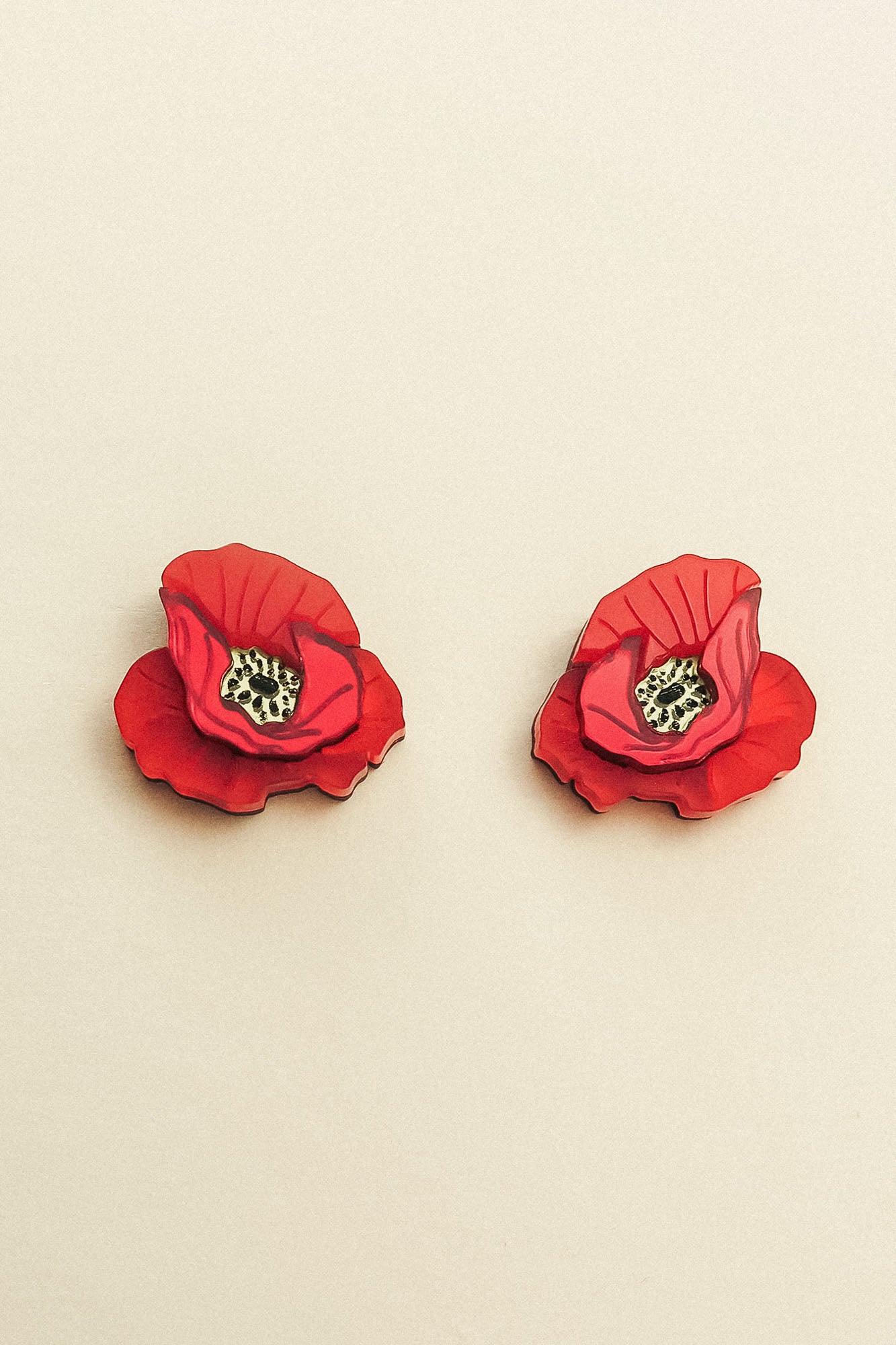 Two red poppy design acrylic earrings with black and white centers are displayed on a neutral beige background, showcasing handcrafted detail.