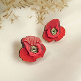 Two red poppy design acrylic earrings with black and yellow centers rest on a white fabric surface. Beige dried foliage in the background creates a soft, elegant setting, highlighting their unique style and lightweight feel.