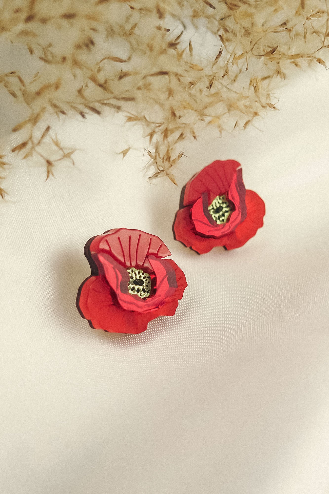 Two red poppy design acrylic earrings with black and yellow centers rest on a white fabric surface. Beige dried foliage in the background creates a soft, elegant setting, highlighting their unique style and lightweight feel.