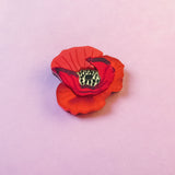 A realistic handmade Red Poppy Flower Lapel Pin with a yellow and black center is placed on a soft, light pink surface, making it a charming accessory for fashion enthusiasts.