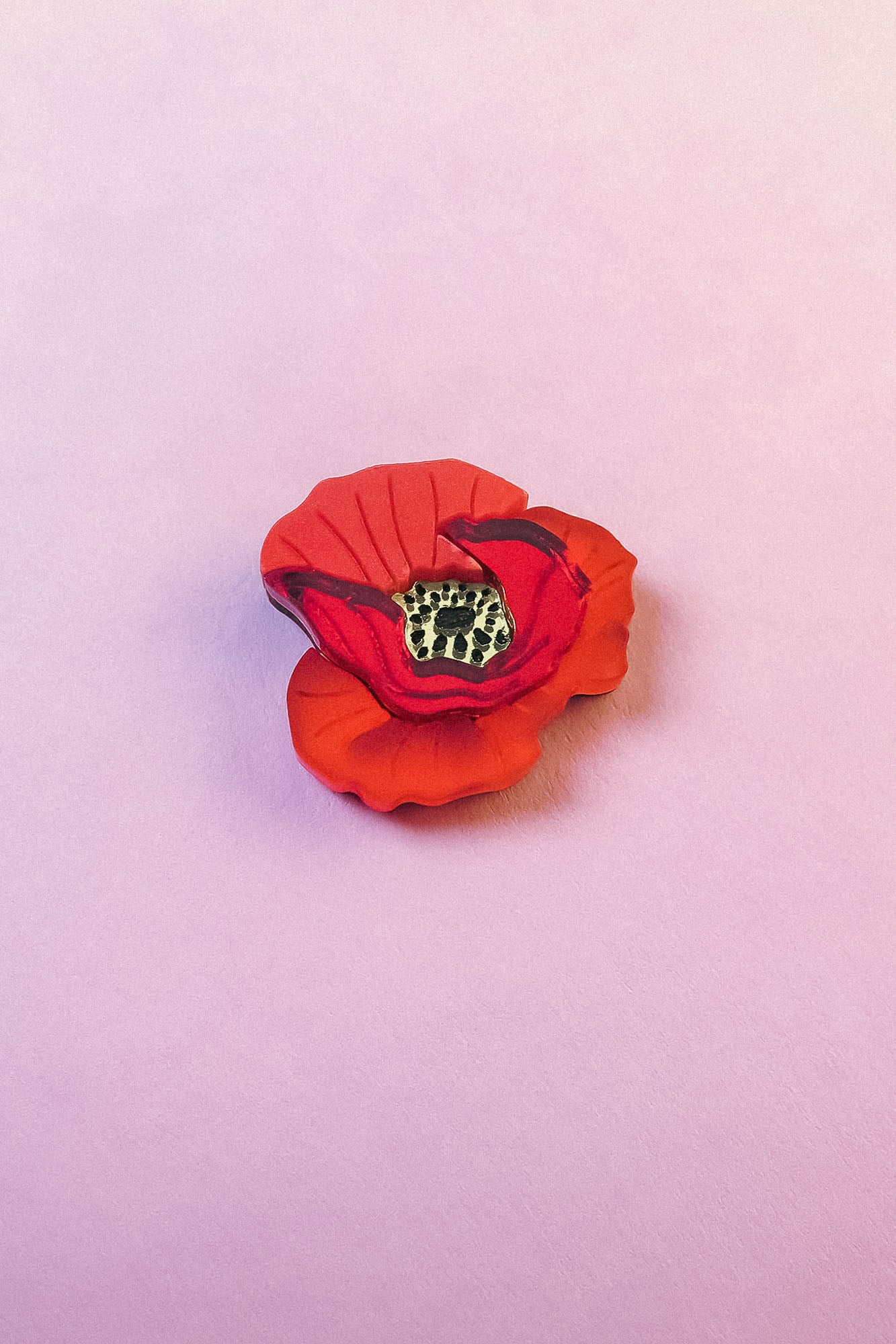 A realistic handmade Red Poppy Flower Lapel Pin with a yellow and black center is placed on a soft, light pink surface, making it a charming accessory for fashion enthusiasts.