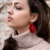 A woman with long brown hair in a beige turtleneck sweater and handcrafted Red Poppy Statement Earrings looks to the side against a blurred outdoor background.