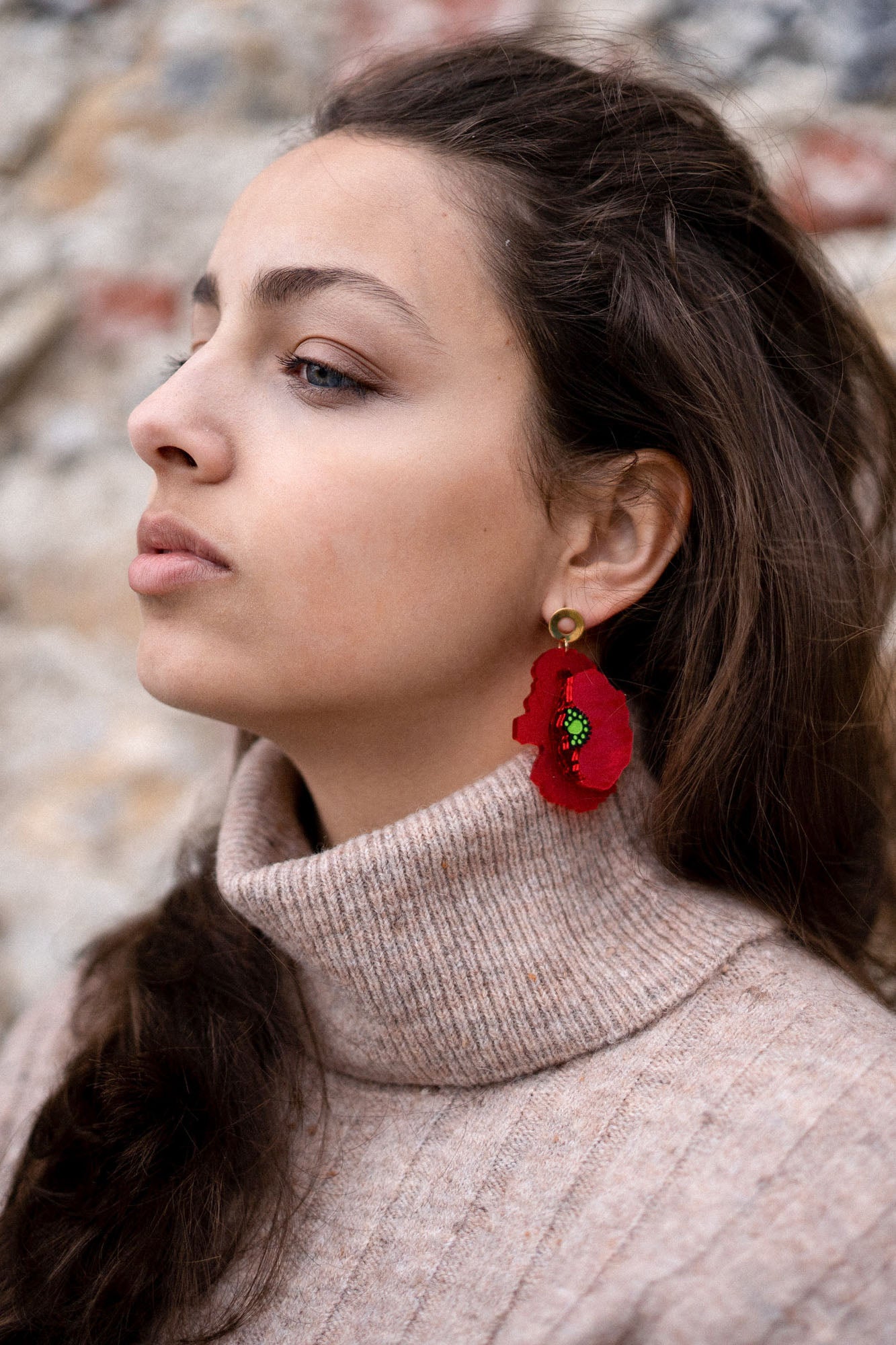 A woman with long brown hair in a beige turtleneck sweater and handcrafted Red Poppy Statement Earrings looks to the side against a blurred outdoor background.