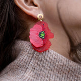 A close-up of a womans ear adorned with handcrafted Red Poppy Statement Earrings—a bold accessory with a green and black center. She has dark hair and is wearing a beige knitted sweater.