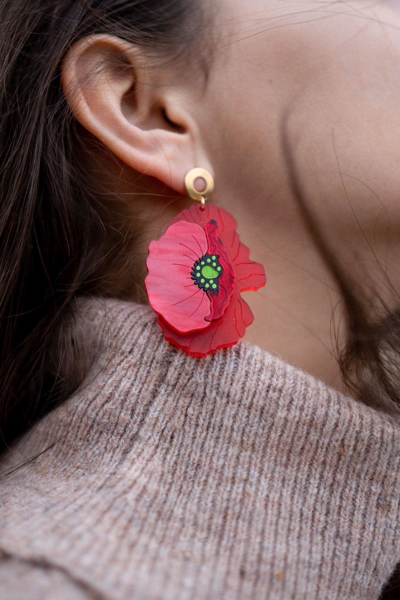 A close-up of a womans ear adorned with handcrafted Red Poppy Statement Earrings—a bold accessory with a green and black center. She has dark hair and is wearing a beige knitted sweater.