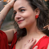 A woman with wavy brown hair, wearing a red dress and lightweight acrylic earrings with a poppy design, stands outdoors among autumn leaves, smiling softly with one hand on her head and the other on her chest.