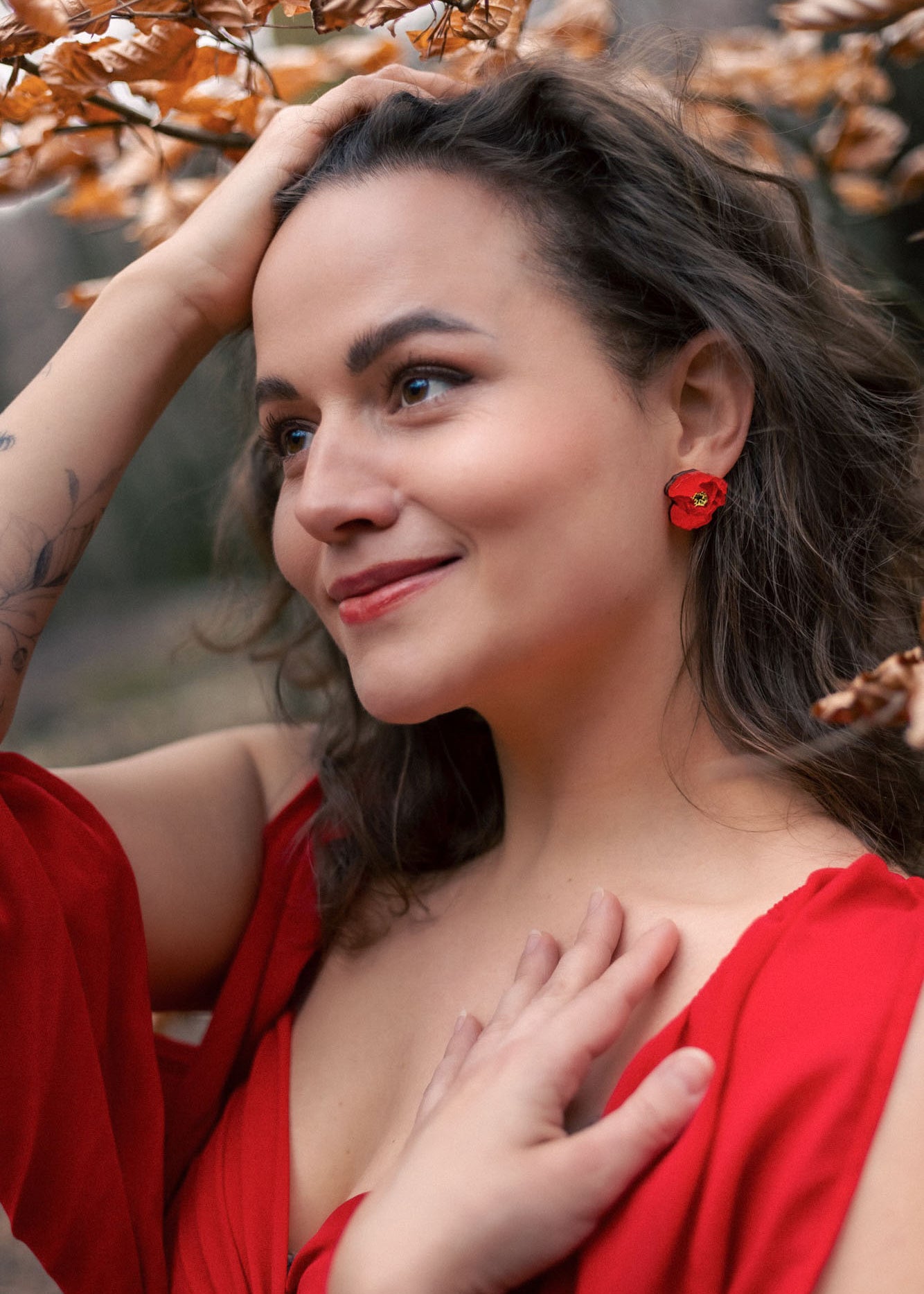 A woman with wavy brown hair, wearing a red dress and lightweight acrylic earrings with a poppy design, stands outdoors among autumn leaves, smiling softly with one hand on her head and the other on her chest.
