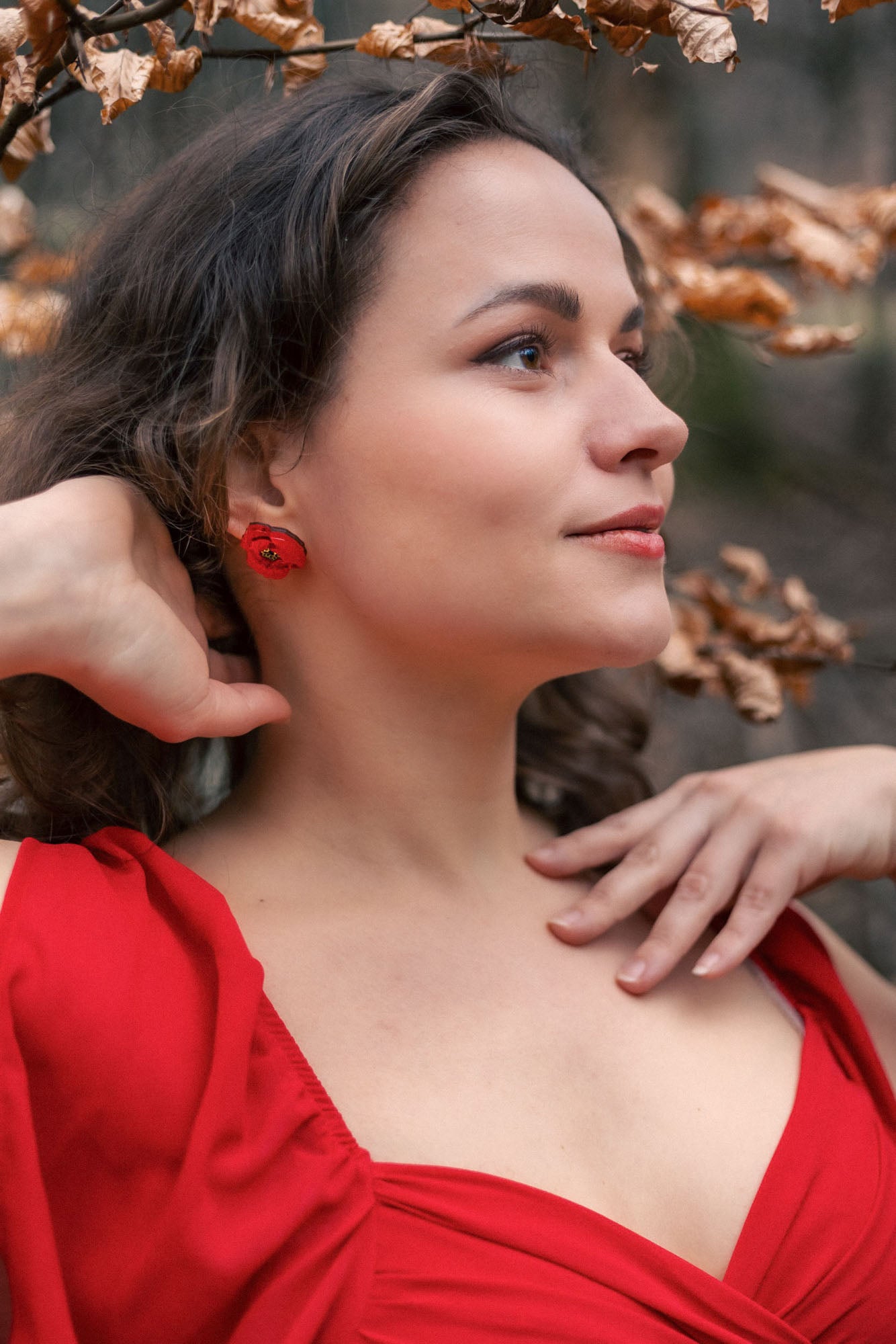 A woman in a red dress poses outdoors near branches with dried leaves. She touches her neck and head, looking to the side with a calm expression. Her wavy brown hair frames lightweight acrylic earrings and a striking red earring.