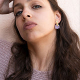 A woman with long brown hair wearing a sparkly pink sweater and handcrafted earrings looks confidently at the camera against a light background. Her hand rests on the side of her head, showing off her elegant accessory.