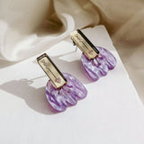 Romantic Tulip Earrings feature rectangular gold-toned tops engraved with hearts and vines, paired with marbled purple acrylic flower-shaped bottoms, offering allergy-free comfort. Displayed on a white surface with a cream fabric background.
