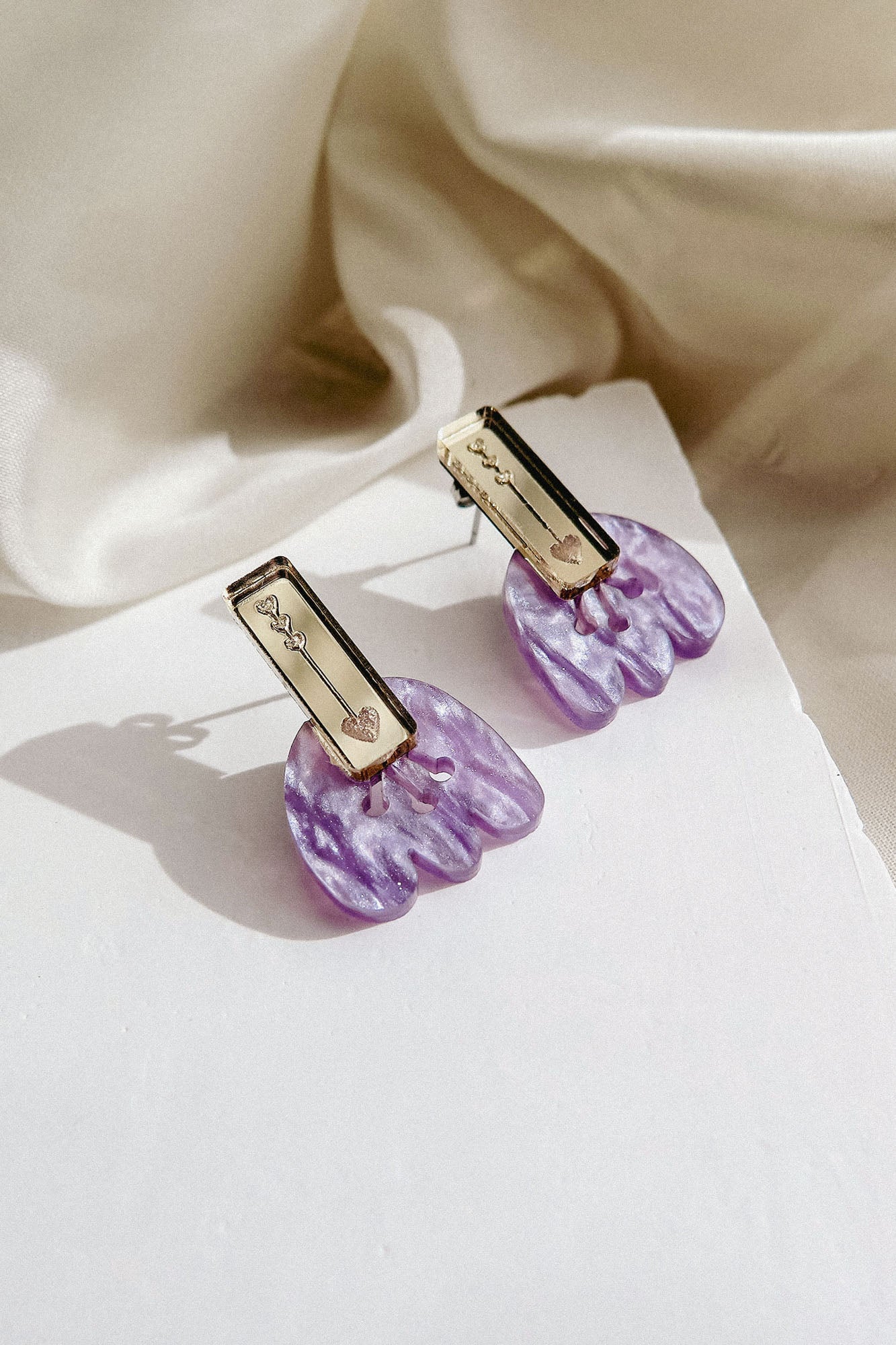 Romantic Tulip Earrings feature rectangular gold-toned tops engraved with hearts and vines, paired with marbled purple acrylic flower-shaped bottoms, offering allergy-free comfort. Displayed on a white surface with a cream fabric background.