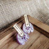 Romantic Tulip Earrings featuring gold rectangular tops engraved with a heart and abstract shapes, attached to marbled purple acrylic flower-shaped bases, offer allergy-free comfort. Displayed on a wooden tray with a burlap background.
