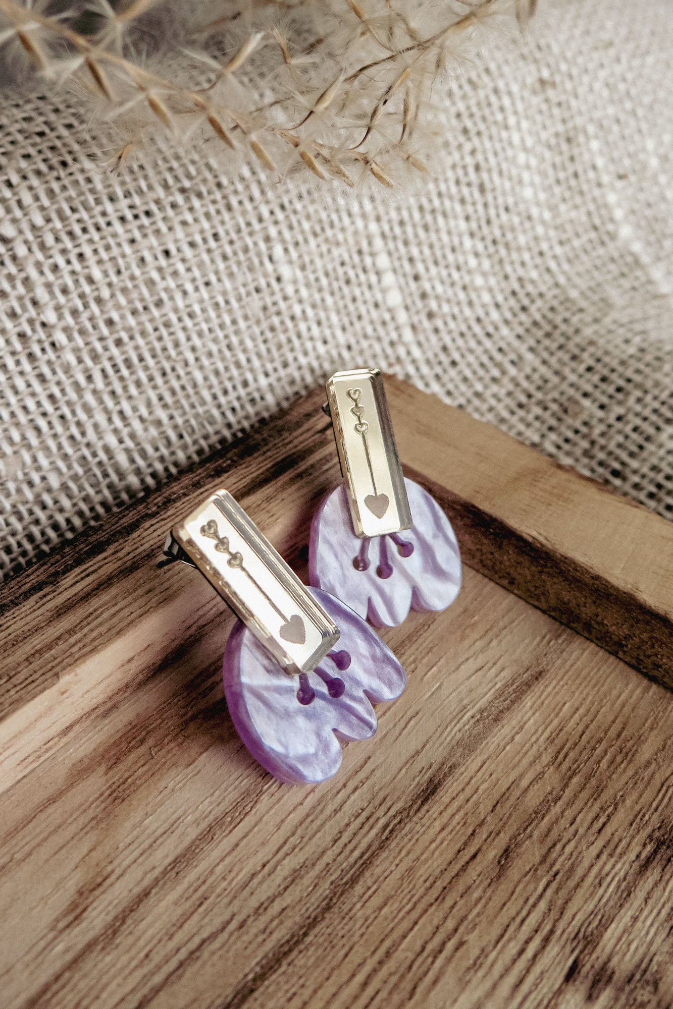 Romantic Tulip Earrings featuring gold rectangular tops engraved with a heart and abstract shapes, attached to marbled purple acrylic flower-shaped bases, offer allergy-free comfort. Displayed on a wooden tray with a burlap background.