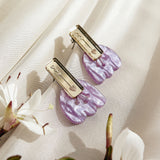 A pair of gold and marbled purple acrylic butterfly-shaped hair clips rest on a soft beige fabric, surrounded by delicate white flowers, offering allergy-free comfort.