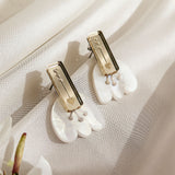 A pair of handcrafted earrings with gold rectangular studs engraved with small designs, attached to white, petal-shaped drops, displayed on a soft, cream-colored fabric with a white flower nearby.