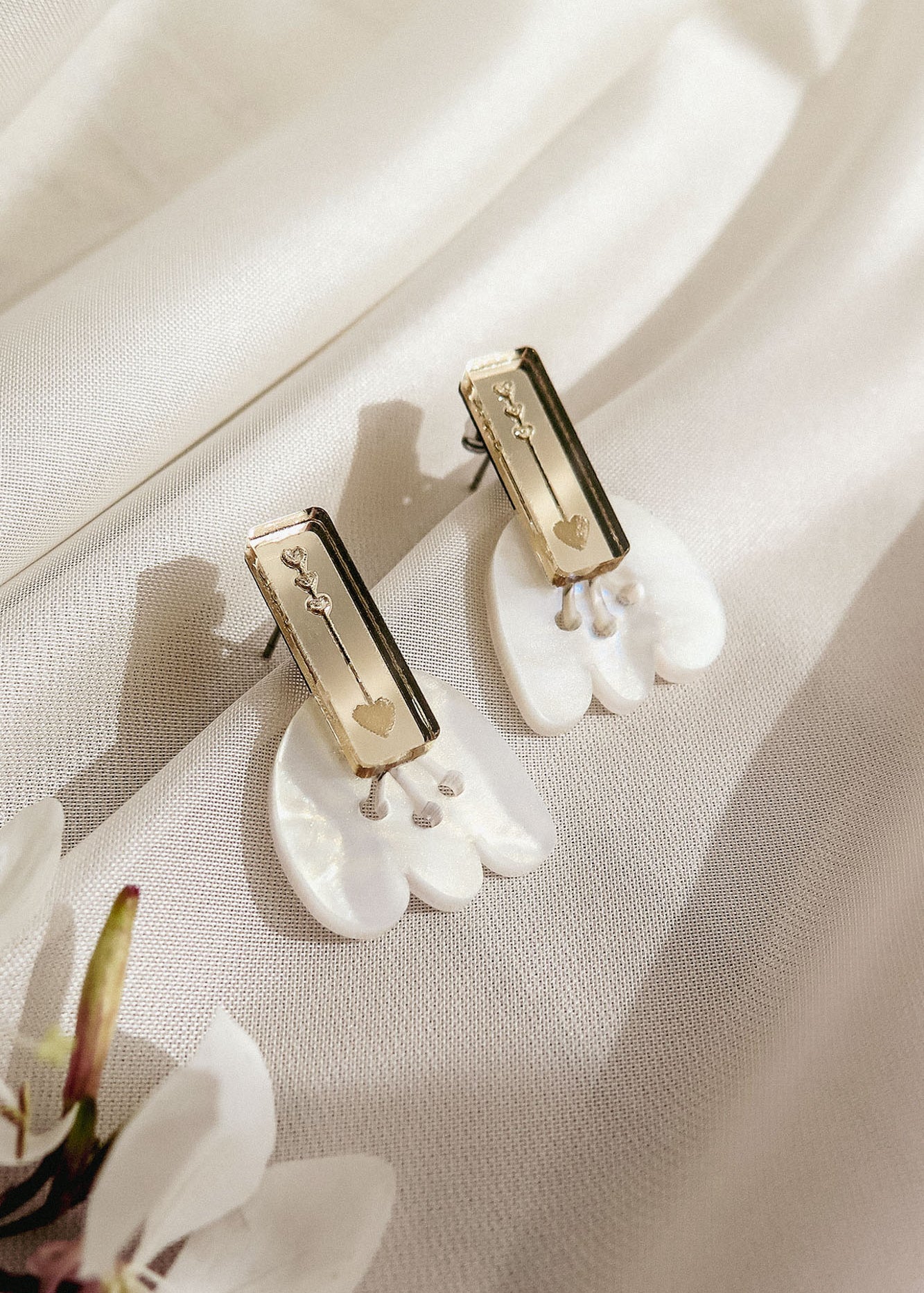 A pair of handcrafted earrings with gold rectangular studs engraved with small designs, attached to white, petal-shaped drops, displayed on a soft, cream-colored fabric with a white flower nearby.
