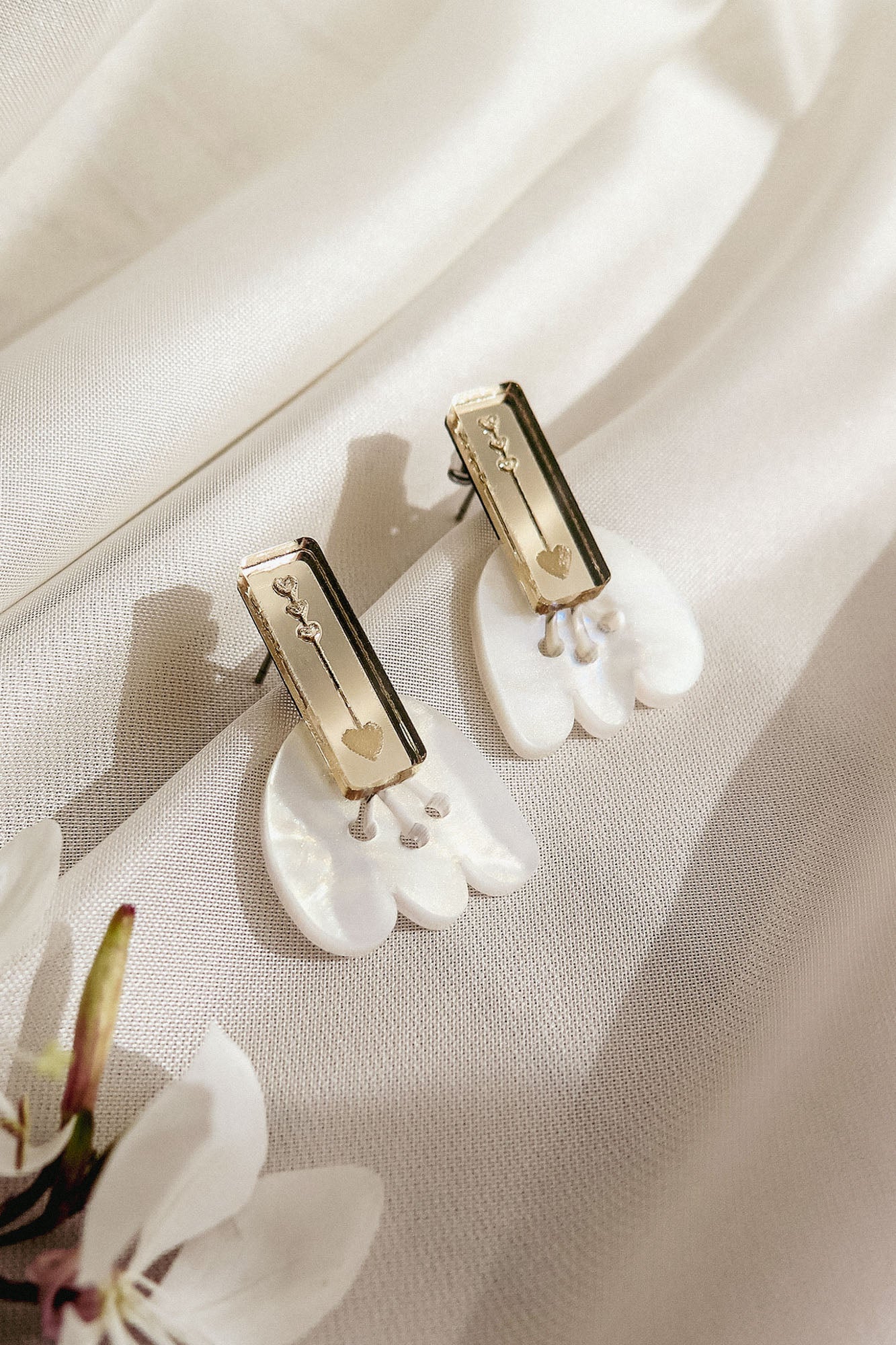 A pair of handcrafted earrings with gold rectangular studs engraved with small designs, attached to white, petal-shaped drops, displayed on a soft, cream-colored fabric with a white flower nearby.
