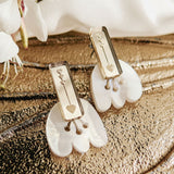 Rectangular gold earrings with engraved key and heart designs rest on white shell-like bases, displayed on a textured metallic surface with white flowers nearby, offering allergy-free comfort.