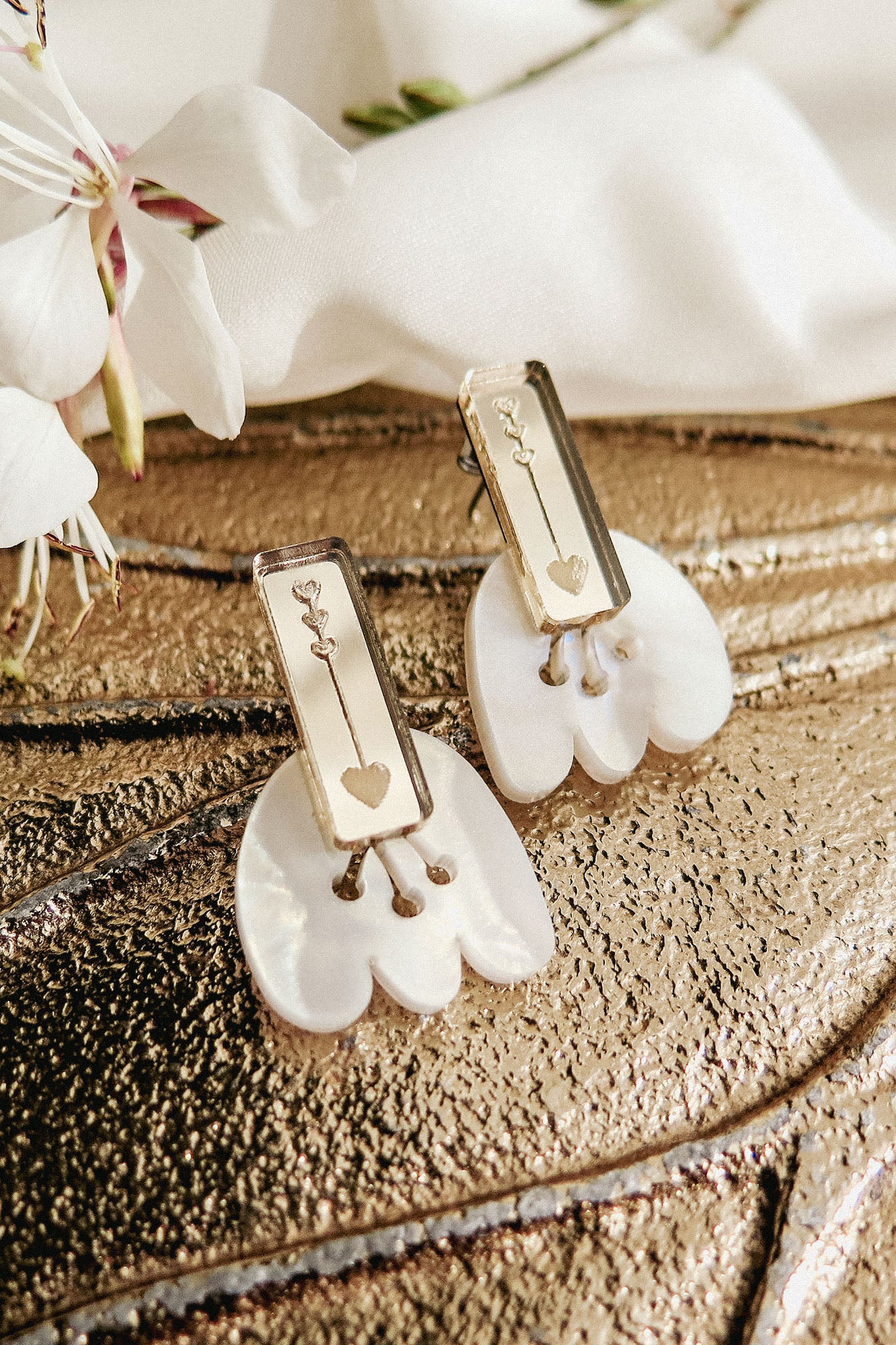 Rectangular gold earrings with engraved key and heart designs rest on white shell-like bases, displayed on a textured metallic surface with white flowers nearby, offering allergy-free comfort.
