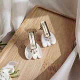 Two pairs of elegant earrings, including Romantic Tulip Earrings with pearl-like white and gold elements, are displayed on a light wooden tray amid soft fabric and blooms, offering allergy-free comfort in a delicate, inviting scene.