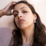 A woman with long brown hair and light eyes gazes confidently at the camera, wearing mauve lipstick, a textured mauve sweater, and Romantic Tulip Earrings in marbled purple acrylic for allergy-free comfort, her hand resting on her head.