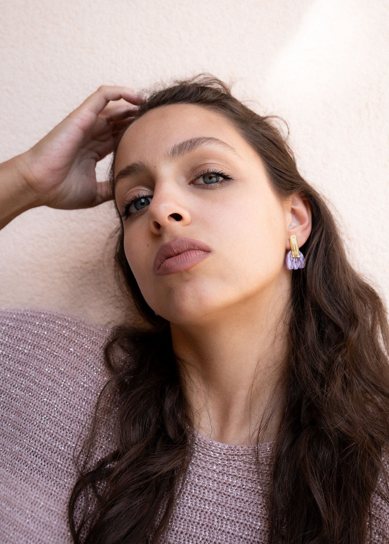 A woman with long brown hair and light eyes gazes confidently at the camera, wearing mauve lipstick, a textured mauve sweater, and Romantic Tulip Earrings in marbled purple acrylic for allergy-free comfort, her hand resting on her head.