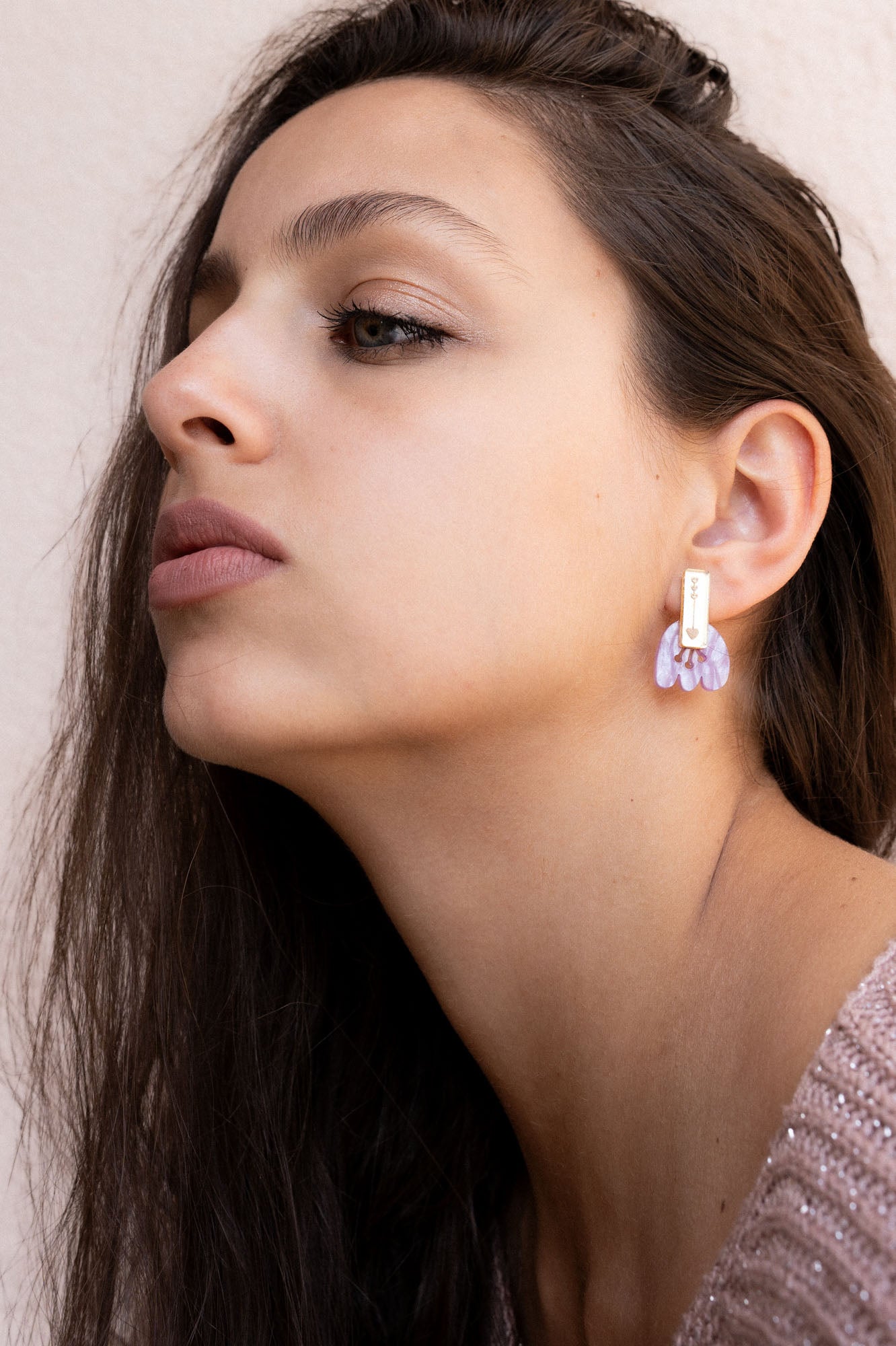 A woman with long brown hair gazes upward, showing off Romantic Tulip Earrings made from marbled purple acrylic. She wears a light pink, textured top, and the background is neutral for allergy-free comfort all day.