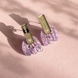A pair of Romantic Tulip Earrings with rectangular gold tops and dangling, marbled purple acrylic flower-shaped pendants, displayed on a soft pink textured surface with delicate shadows for allergy-free comfort.