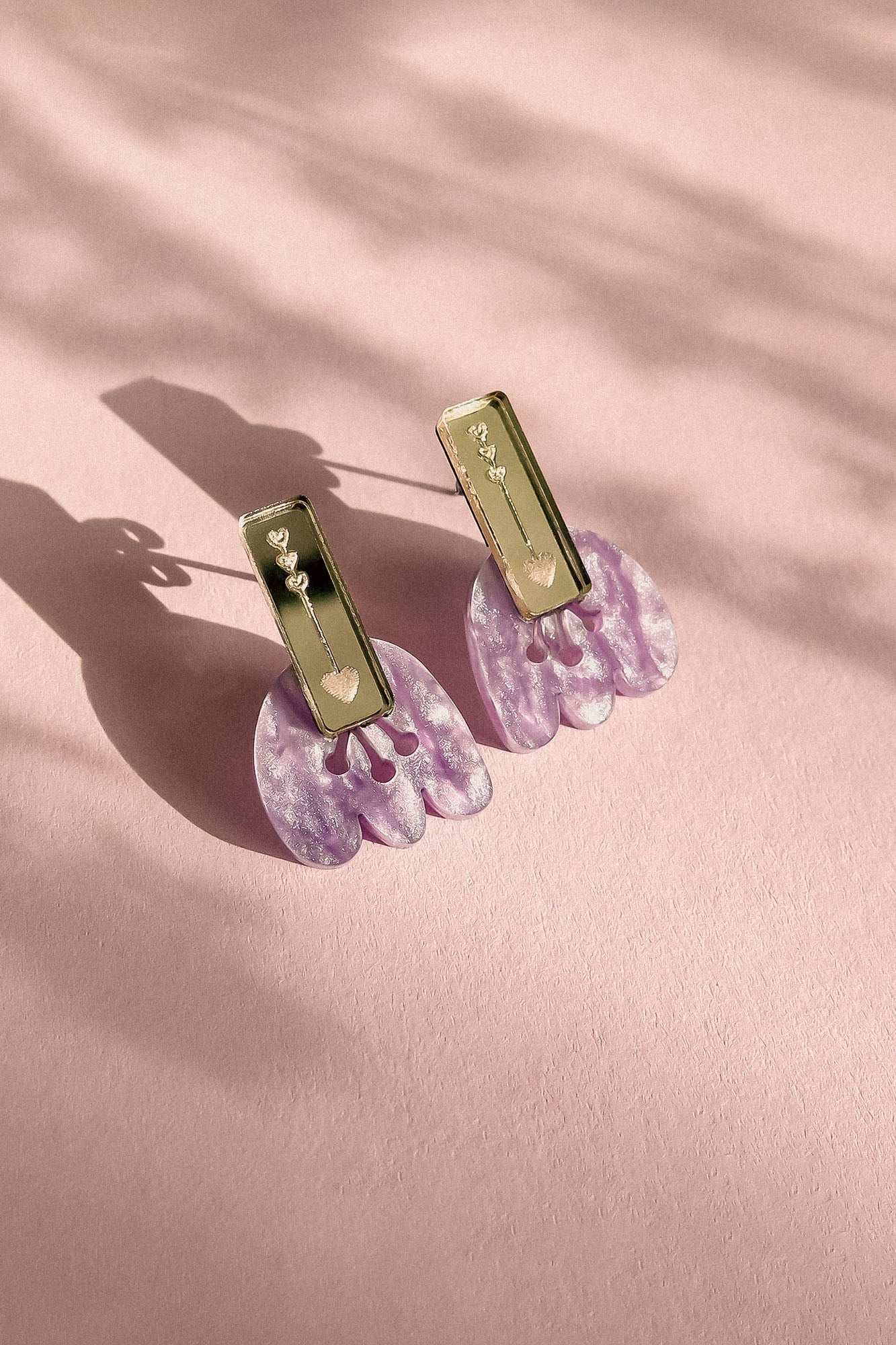 A pair of Romantic Tulip Earrings with rectangular gold tops and dangling, marbled purple acrylic flower-shaped pendants, displayed on a soft pink textured surface with delicate shadows for allergy-free comfort.