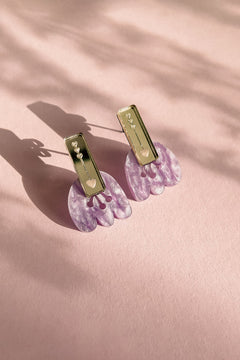 A pair of Romantic Tulip Earrings with rectangular gold tops and dangling, marbled purple acrylic flower-shaped pendants, displayed on a soft pink textured surface with delicate shadows for allergy-free comfort.