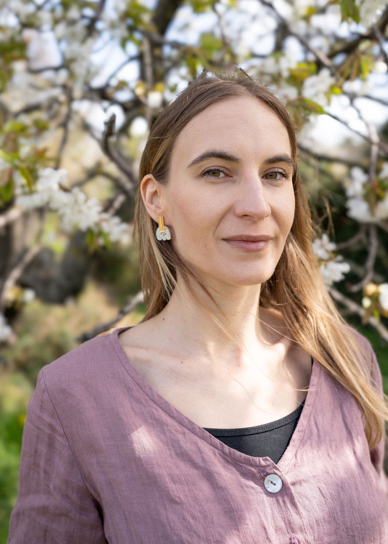 A woman with long blonde hair, wearing a mauve top and Romantic Tulip Earrings in marbled purple acrylic, stands outdoors in front of a blooming tree with white flowers. Sunlight highlights her face and the spring blossoms behind her.