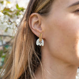 Close-up of a woman wearing Romantic Tulip Earrings in gold and white, with her light brown hair tucked behind her ear. Enjoy allergy-free comfort as blurred greenery and white flowers create a soft, romantic backdrop.