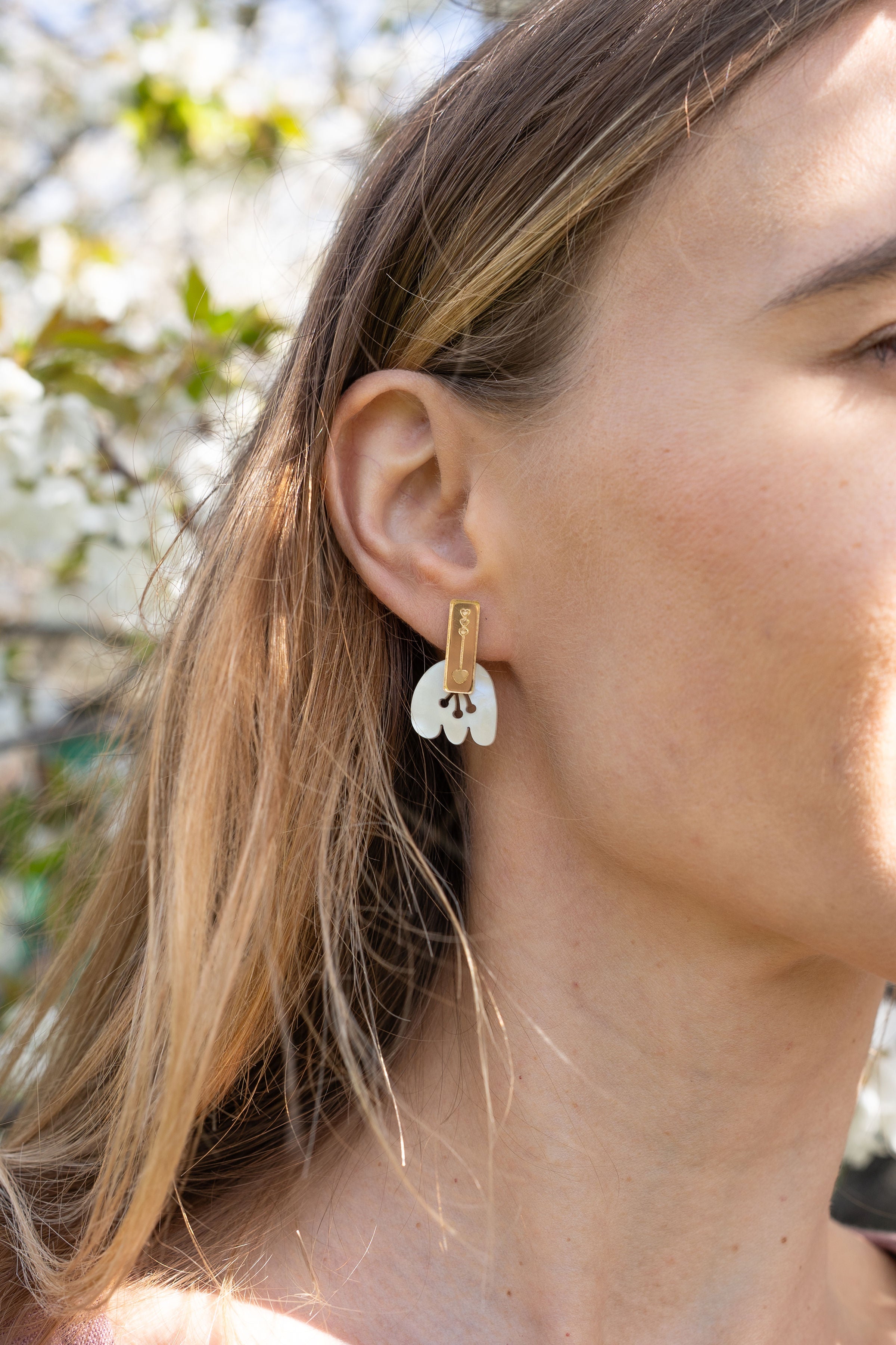 Close-up of a woman wearing Romantic Tulip Earrings in gold and white, with her light brown hair tucked behind her ear. Enjoy allergy-free comfort as blurred greenery and white flowers create a soft, romantic backdrop.