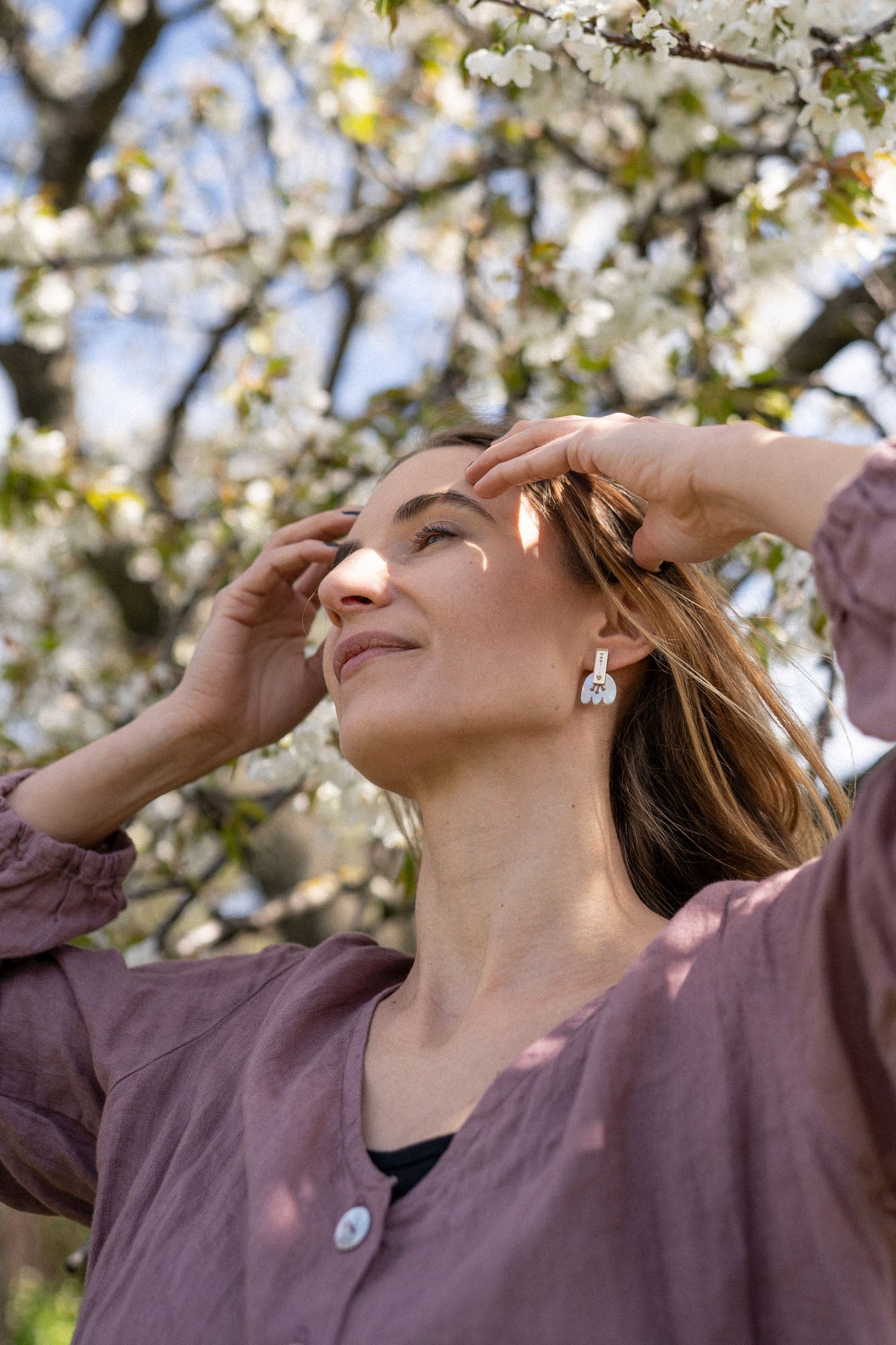 A woman wearing a mauve blouse and Romantic Tulip Earrings stands outside with her hands near her face, eyes closed, enjoying sunlight beneath blossoming tree branches. The marbled purple acrylic adds an allergy-free comfort to her serene moment.