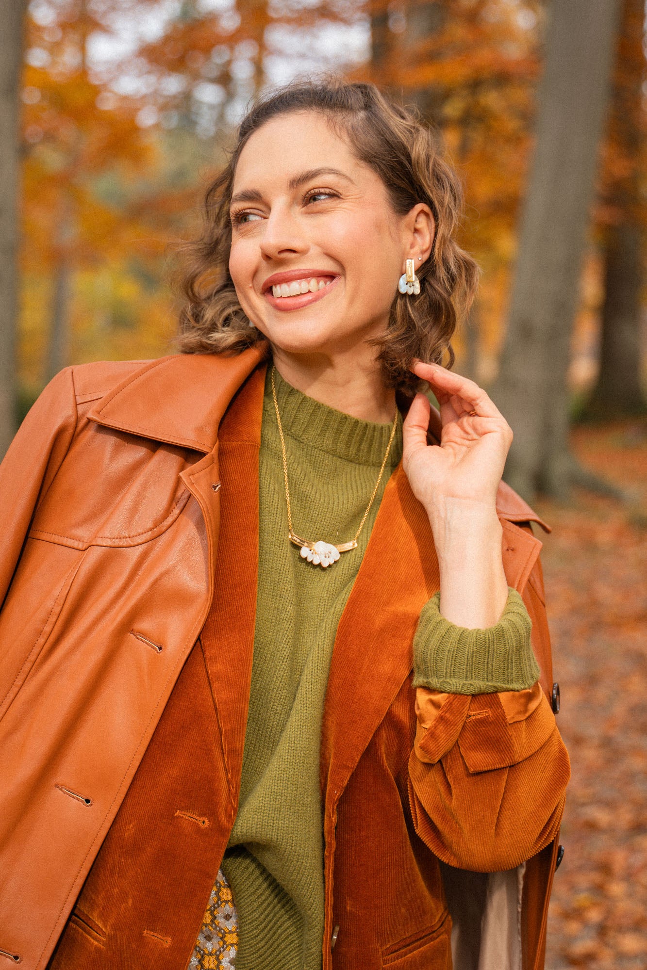 A woman smiles elegantly while standing in a forest with autumn leaves. She wears a brown leather coat over an orange suede jacket, with a green sweater, patterned skirt, and a handcrafted Tulip Necklace. Tall trees and fallen leaves fill the background.