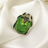 A handcrafted accessory, this green Rose Chafer Pin with black legs and silver accents rests on smooth white satin fabric, a few dried plant stems in the background highlighting its meaningful design.