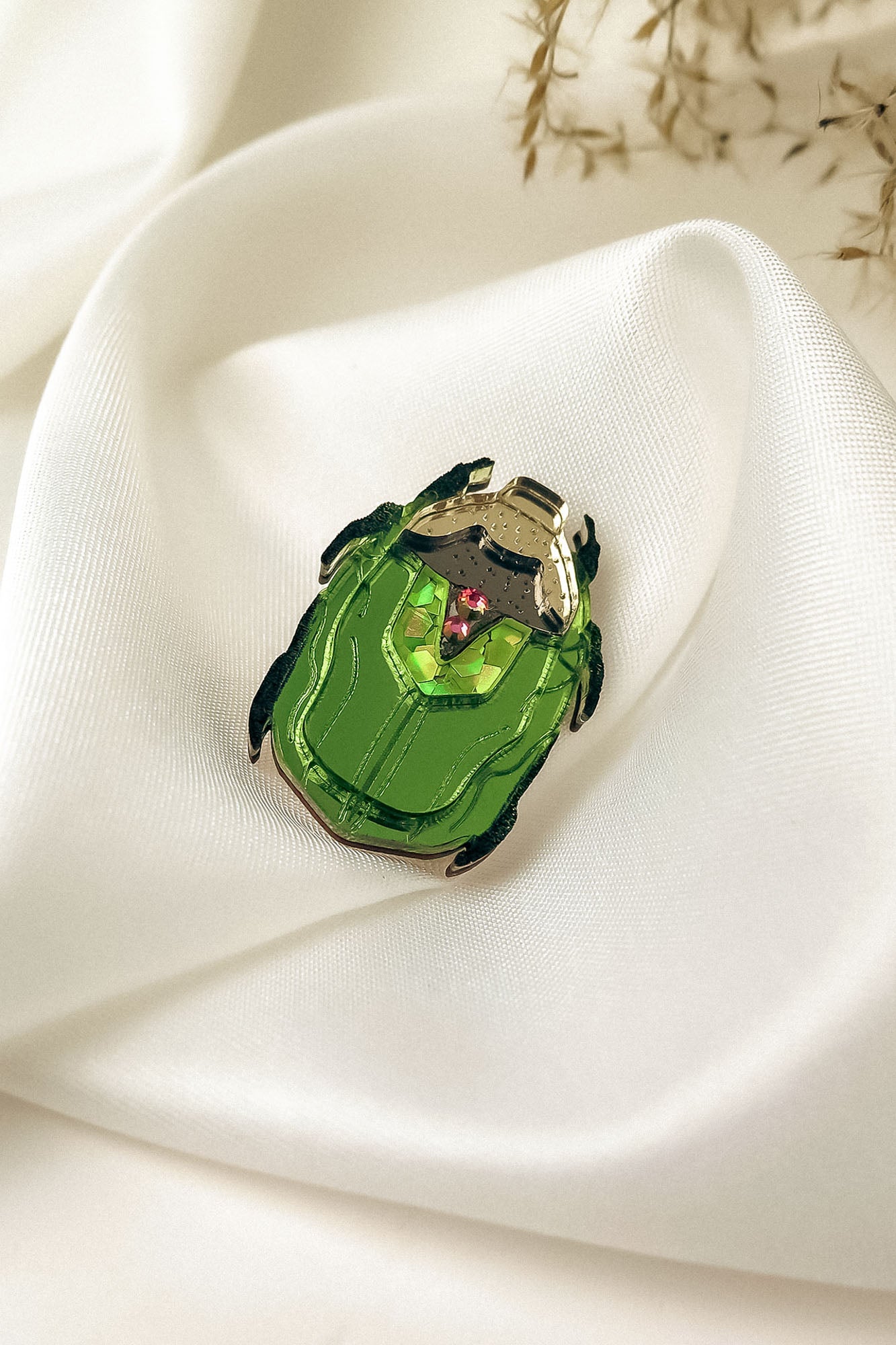 A handcrafted accessory, this green Rose Chafer Pin with black legs and silver accents rests on smooth white satin fabric, a few dried plant stems in the background highlighting its meaningful design.