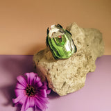 A green and white Rose Chafer Pin brooch with black accents rests on a light-colored rock next to a purple flower, all placed on a pastel purple surface with a beige background—an elegant, handcrafted accessory with meaningful design.