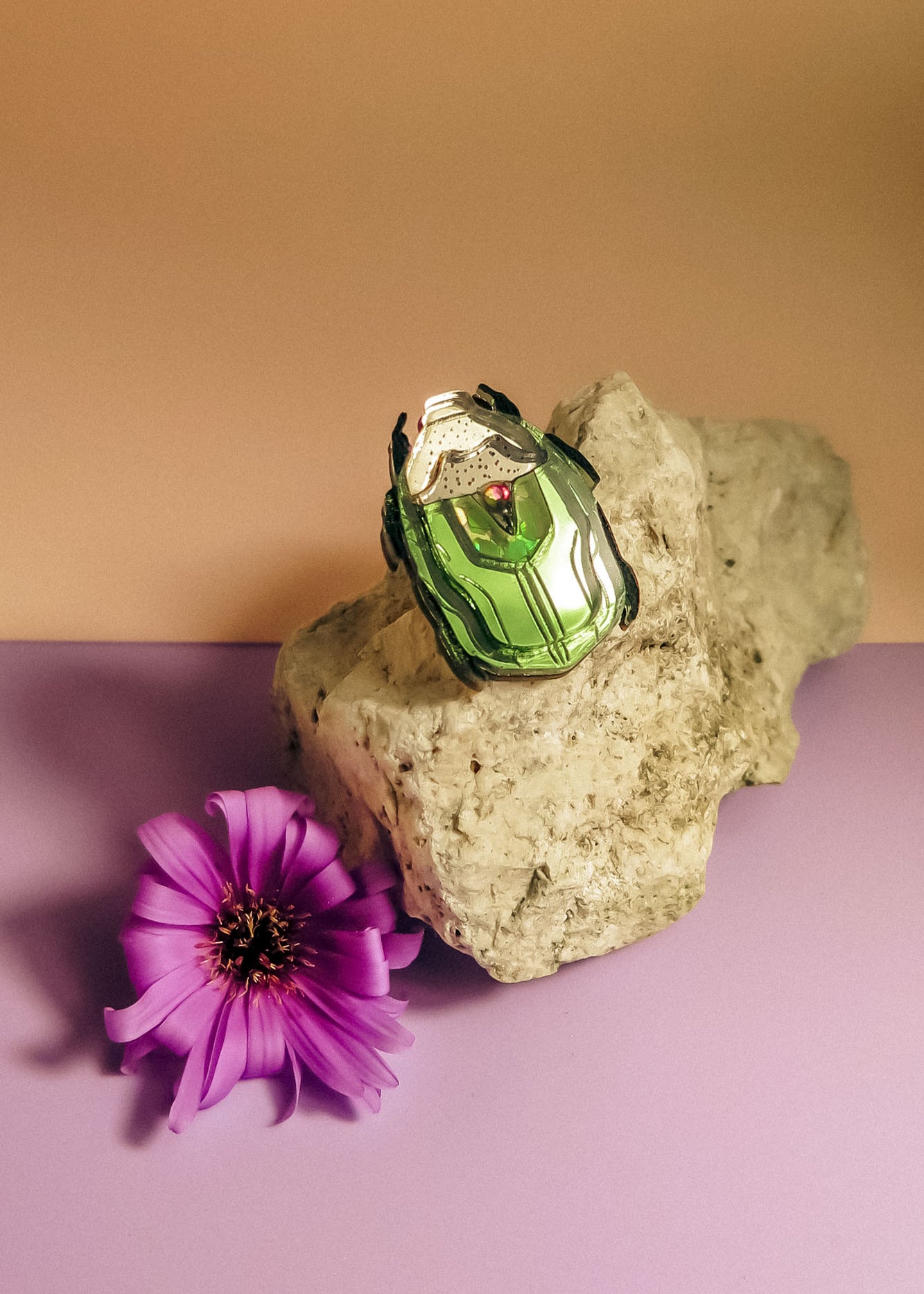 A green and white Rose Chafer Pin brooch with black accents rests on a light-colored rock next to a purple flower, all placed on a pastel purple surface with a beige background—an elegant, handcrafted accessory with meaningful design.