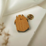 A wooden pin with a metal backing rests on a white surface near a piece of white fabric. This handcrafted accessory features an abstract, organic shape, and a gold-colored clasp is placed nearby.