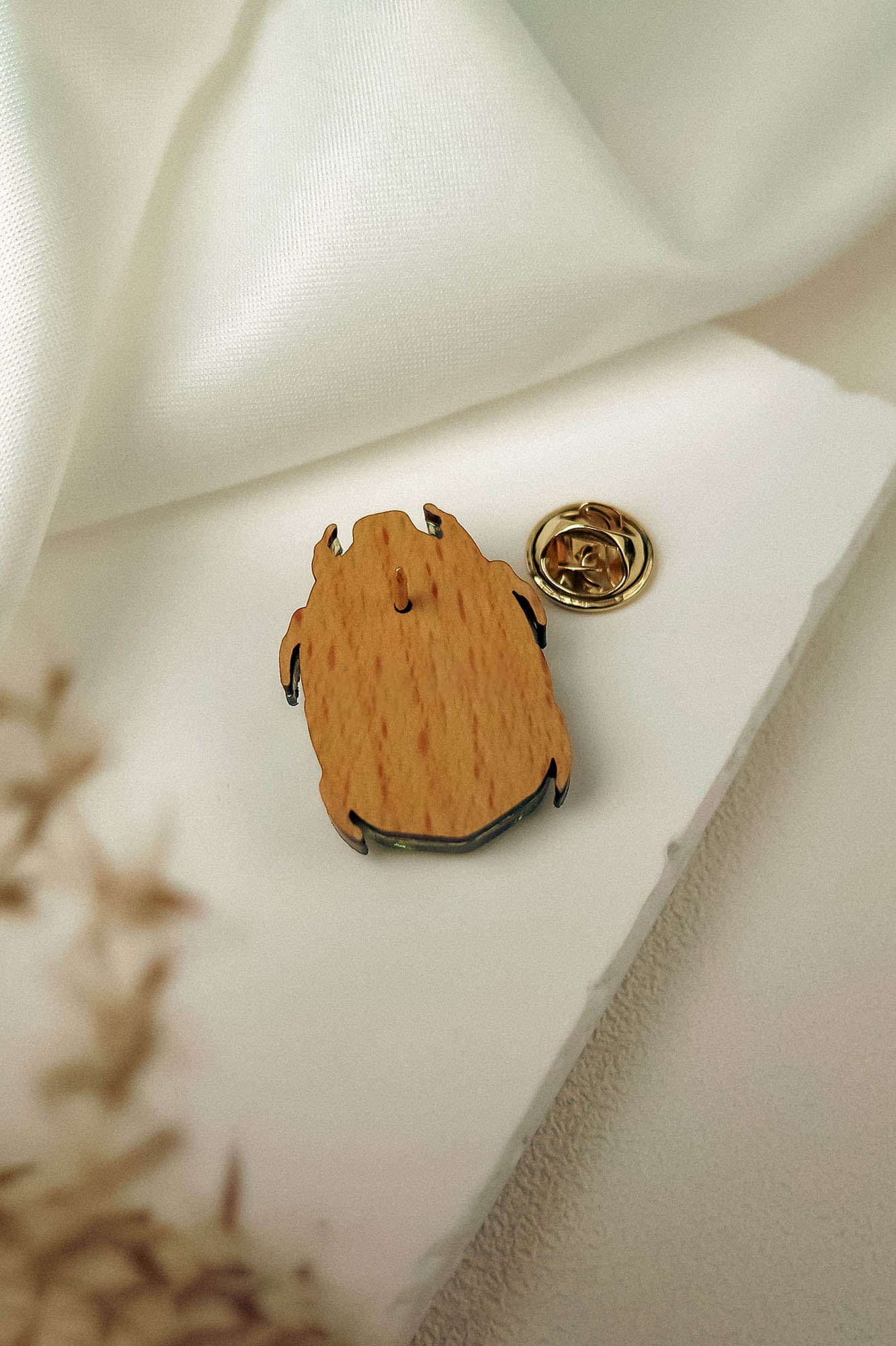 A wooden pin with a metal backing rests on a white surface near a piece of white fabric. This handcrafted accessory features an abstract, organic shape, and a gold-colored clasp is placed nearby.