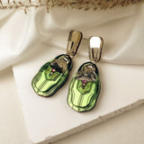 A pair of elegant, nature-inspired Rose Chafer Earrings with green, gem-like pendants and metallic gold hooks, displayed on a white textured surface with fabric and dried plant accents.