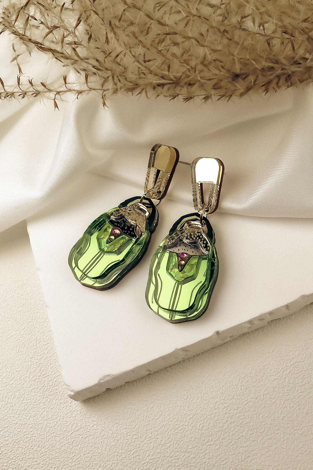 A pair of elegant, nature-inspired Rose Chafer Earrings with green, gem-like pendants and metallic gold hooks, displayed on a white textured surface with fabric and dried plant accents.