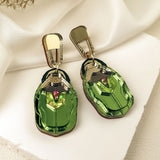 A pair of metallic green, nature-inspired Rose Chafer Earrings with car shapes and gold-colored clips, displayed on a white textured surface with soft white fabric and dried flowers in the background.