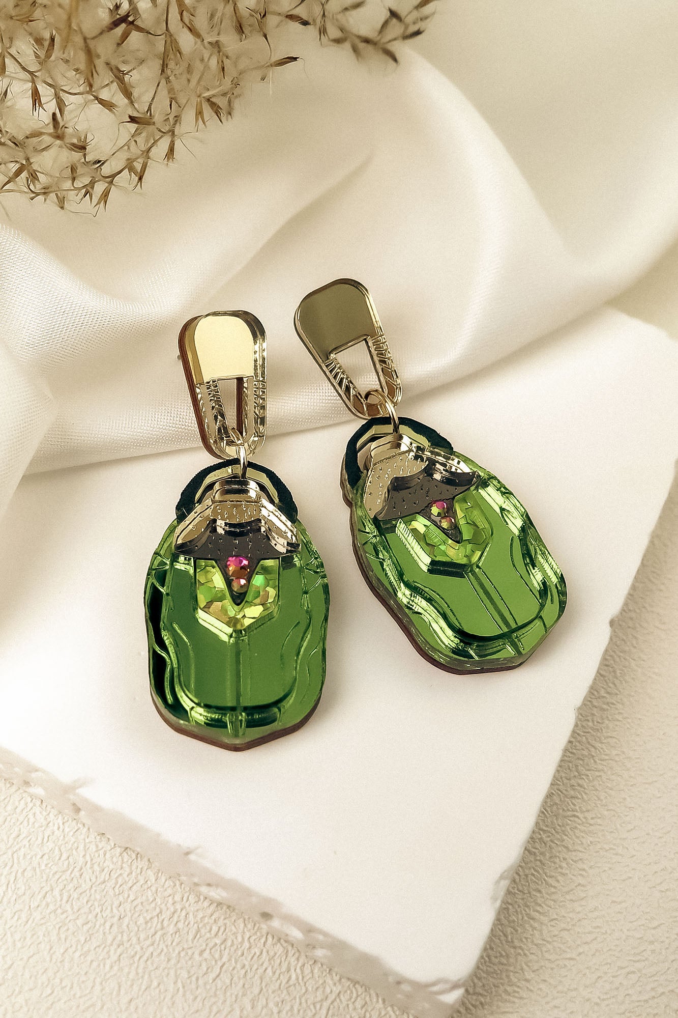 A pair of metallic green, nature-inspired Rose Chafer Earrings with car shapes and gold-colored clips, displayed on a white textured surface with soft white fabric and dried flowers in the background.