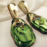 A close-up of handcrafted Rose Chafer Earrings, featuring gold and green geometric shapes with a shiny metallic finish, placed on a soft white fabric background for a nature-inspired look.