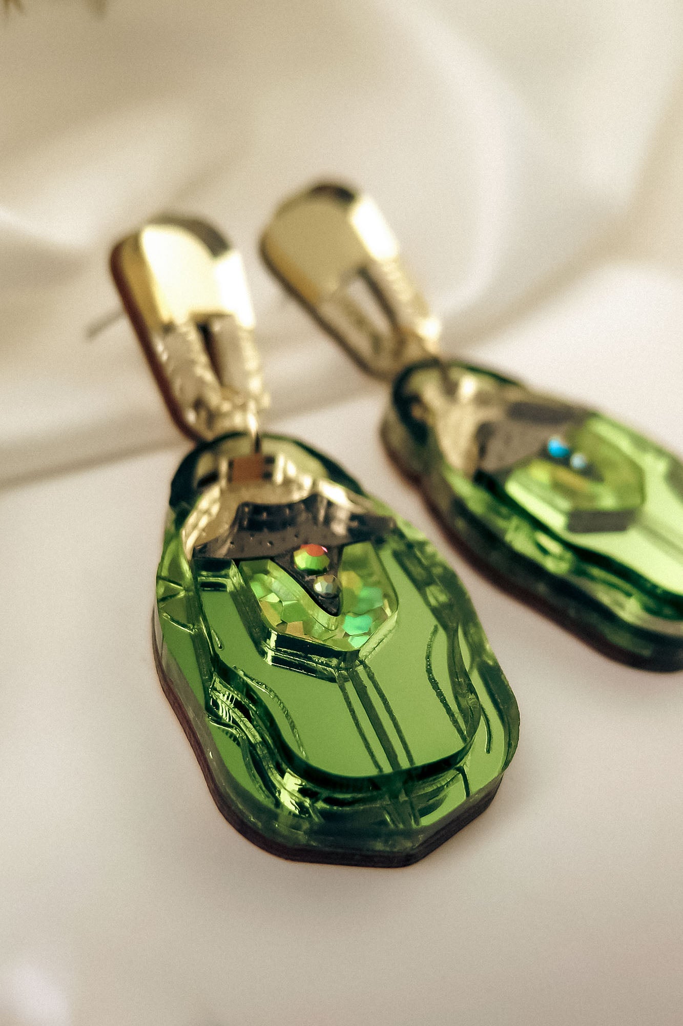 A close-up of handcrafted Rose Chafer Earrings, featuring gold and green geometric shapes with a shiny metallic finish, placed on a soft white fabric background for a nature-inspired look.