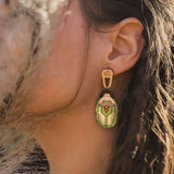 A close-up of a womans ear adorned with Rose Chafer Earrings, handcrafted and nature-inspired, featuring an illustrated figure in green clothing. Her brown hair partially covers her ear while dried grass adds texture to the foreground.