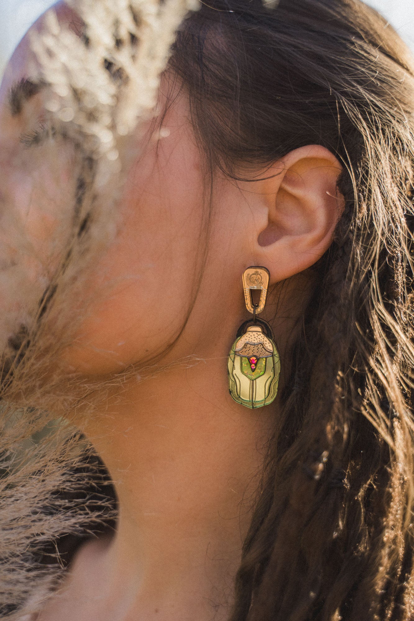 A close-up of a womans ear adorned with Rose Chafer Earrings, handcrafted and nature-inspired, featuring an illustrated figure in green clothing. Her brown hair partially covers her ear while dried grass adds texture to the foreground.