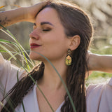 A woman with long, braided hair and bold Rose Chafer Earrings stands outdoors with her eyes closed, smiling softly and raising her arms behind her head in a relaxed, nature-inspired setting among tall grass.